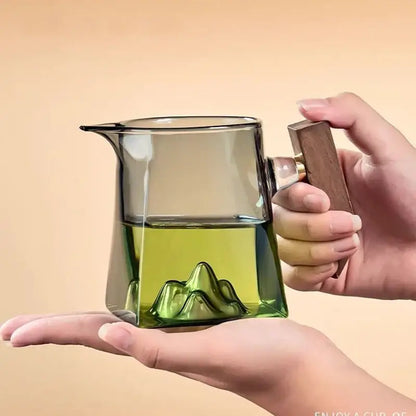 A clear glass tea cup set with wooden handle featuring a green mountain design filled with light green tea