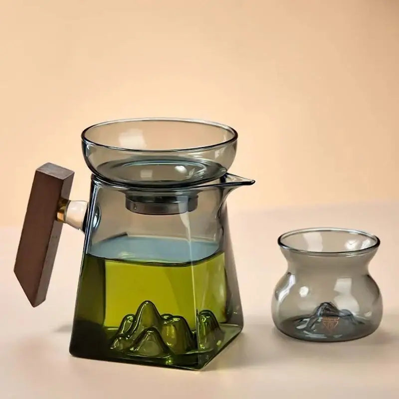 A sleek transparent glass tea cup set with wooden handle and mountain-shaped base filled with green tea