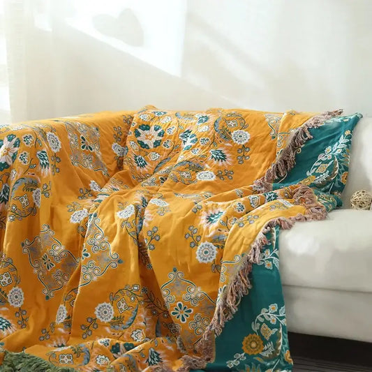 A mustard yellow cotton throw blanket with teal and white decorbites gauze flowers and a soft fringed edge
