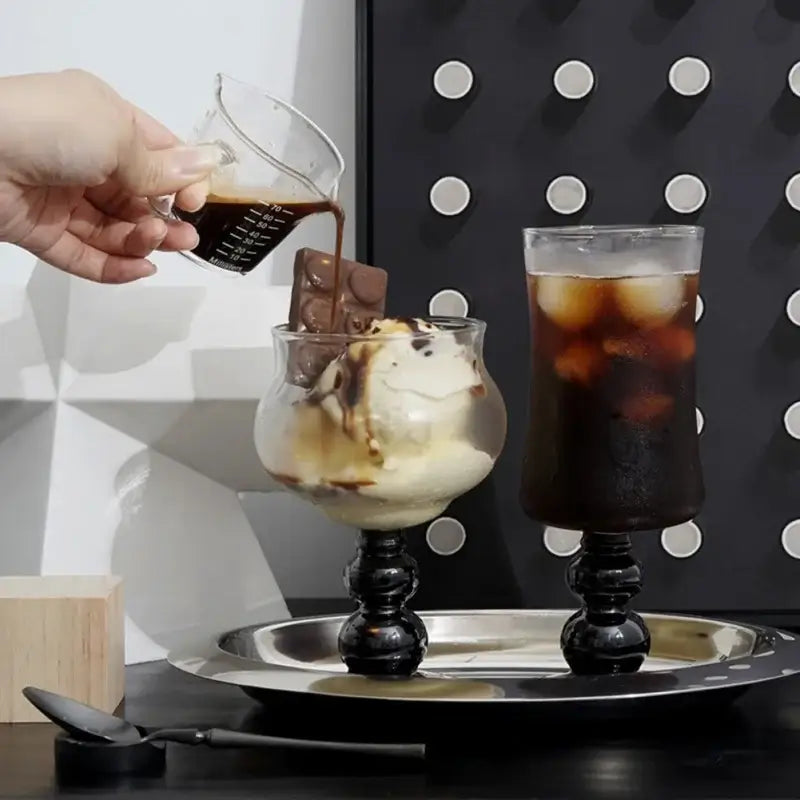 A clear glass dessert cup with black ornate decorbites glass beaded stem filled with vanilla ice cream and chocolate sauce
