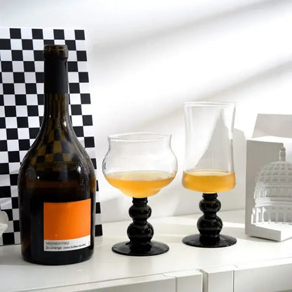 A dark amber wine bottle with an orange label beside two black-stemmed nordic black glass beaded stem goblets filled with golden liquid