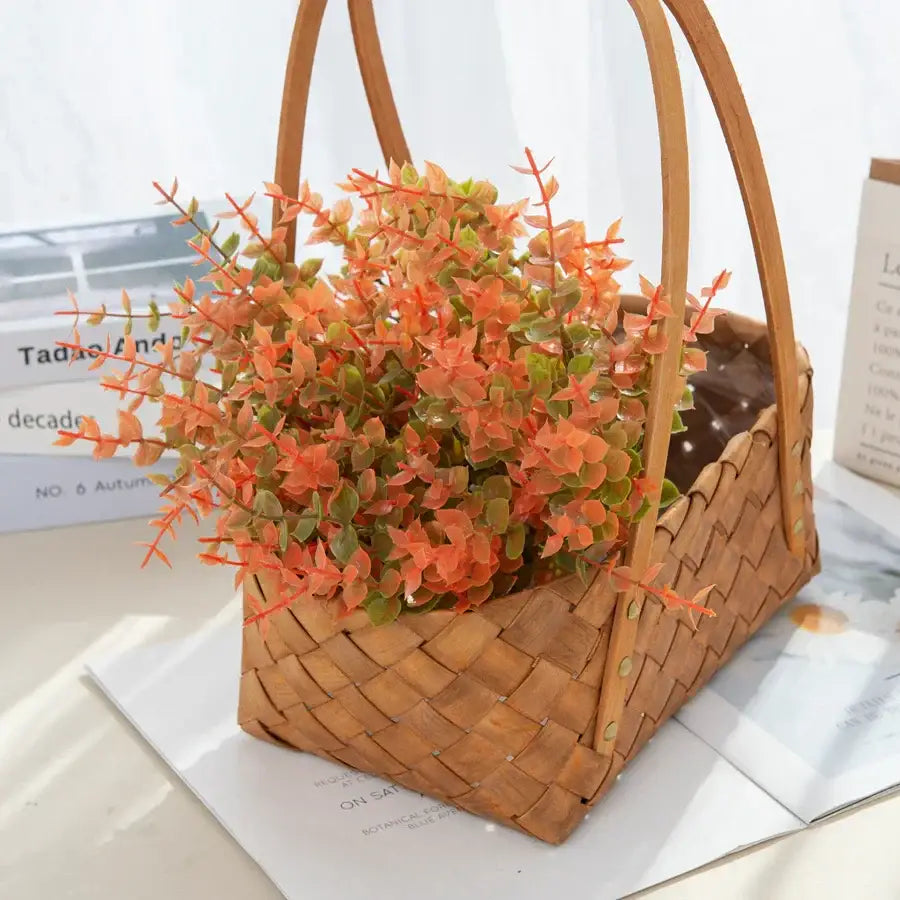 A woven bamboo basket with curved handles filled with artificial orange and green eucalyptus leaves for garden decor