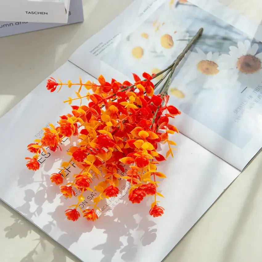 Orange and red artificial eucalyptus leaves arranged with vibrant clustered blossoms in white pitcher for garden decor