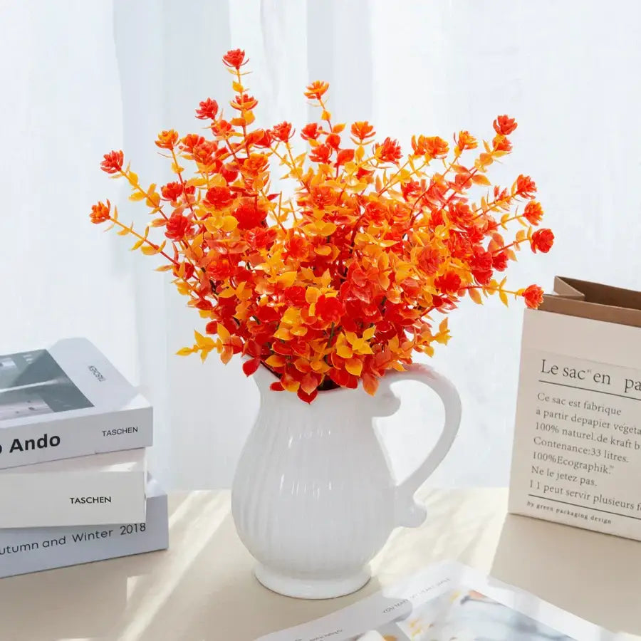 A vibrant bouquet of artificial eucalyptus leaves in orange and red arranged in a white ceramic pitcher for garden decor