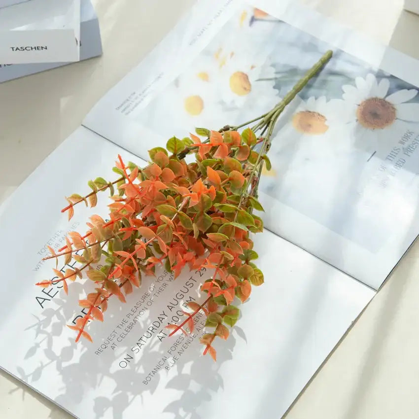 A vibrant orange and green artificial eucalyptus leaves arrangement with detailed leaves in a white pitcher garden decor