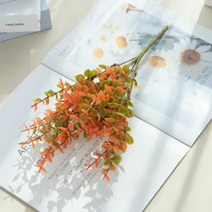 A vibrant orange and green artificial eucalyptus leaves arrangement with detailed leaves in a white pitcher garden decor