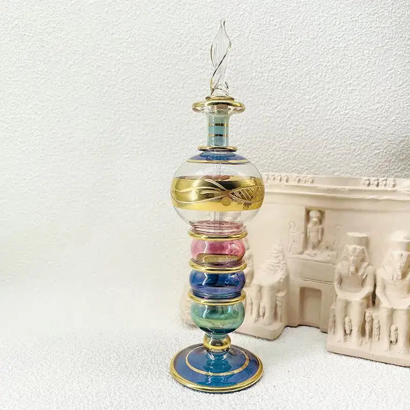A tall ornate glass bottle jewelry ornament with twisted gold cap and layered pink, blue, green, and teal sections