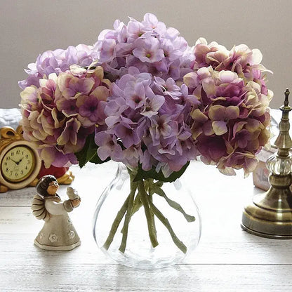 A clear glass vase holding a pastel decorbites hydrangea branch silk flower bouquet with soft purple and cream petals