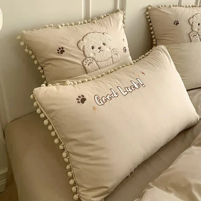 Cream-colored cotton bedding set pillow with embroidered cartoon bear applique and good luck text trimmed in pom-poms