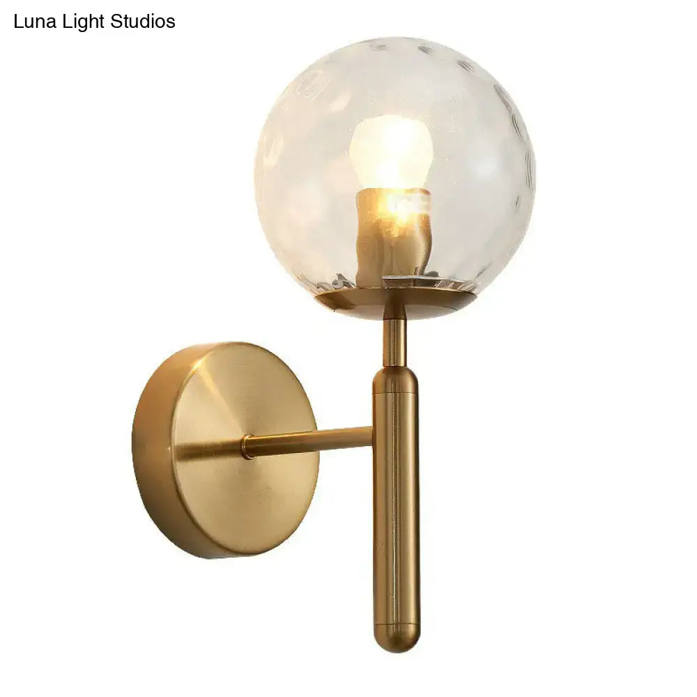 A modern brass-finished wall sconce with a textured, clear glass globe housing an illuminated bulb.