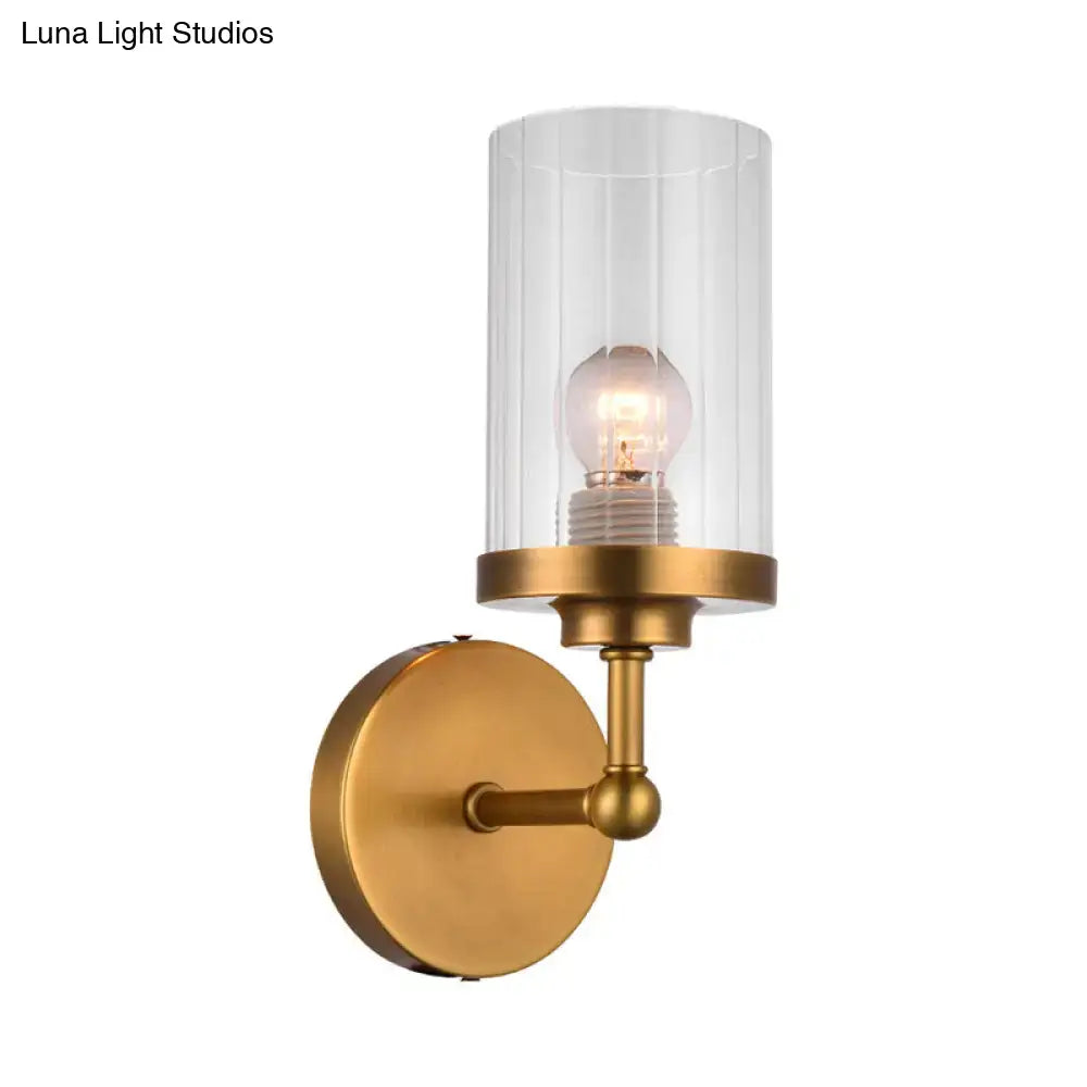 A sleek postmodern brass wall light with a clear cylindrical glass shade and exposed bulb for bedroom use