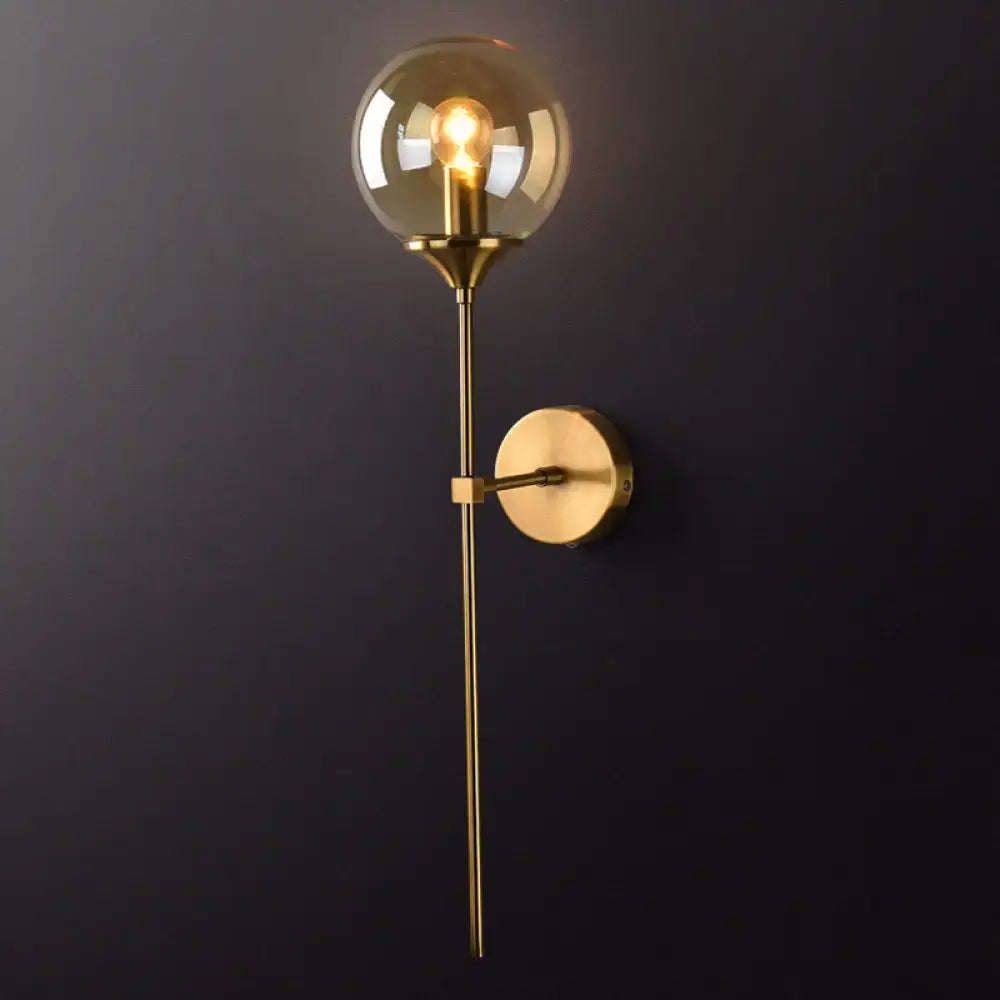 Postmodern brass pencil arm wall lamp with gold-toned stem and spherical amber glass shade for bedroom sconce use