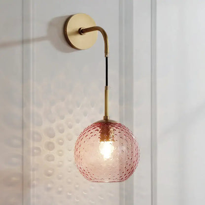 A pink glass globe wall lamp with hammered texture suspended by brass arm and black cord in pink yellow blue style