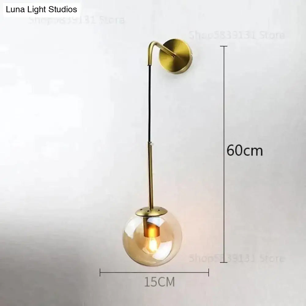 A modern brass-finished wall lamp with a spherical amber glass globe and curved arm, ideal for bedroom lighting