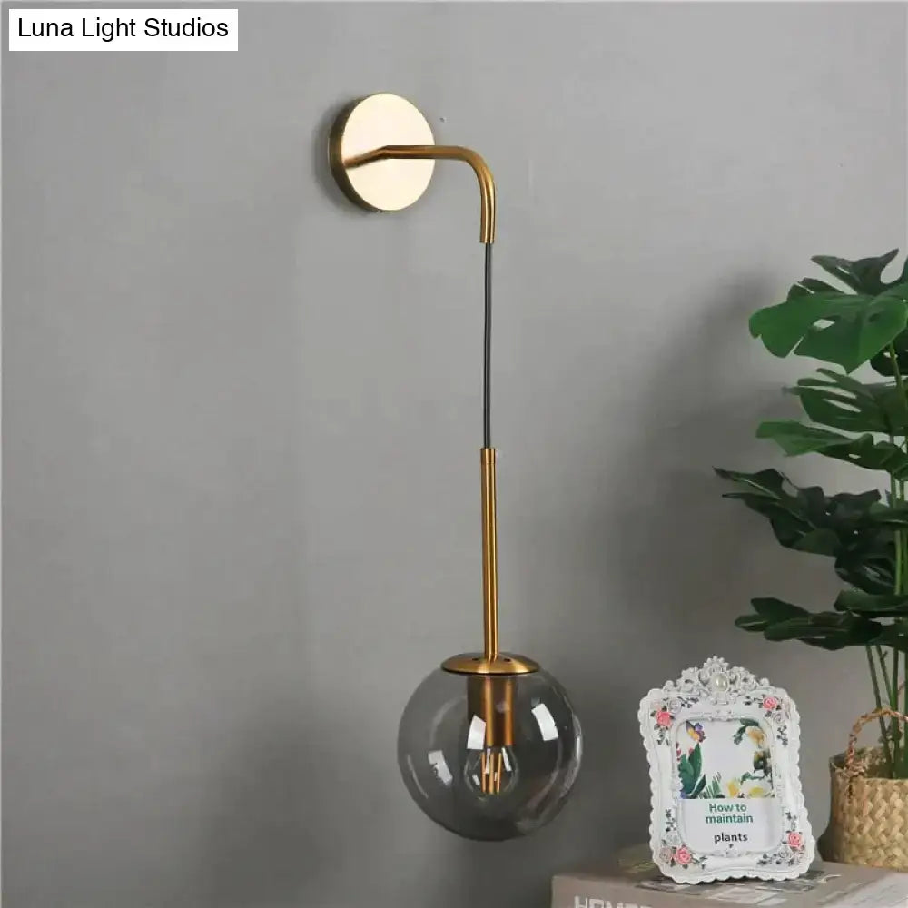 A sleek brass-finished nordic retro modern wall lamp with a curved arm and clear spherical glass shade for bedroom lighting
