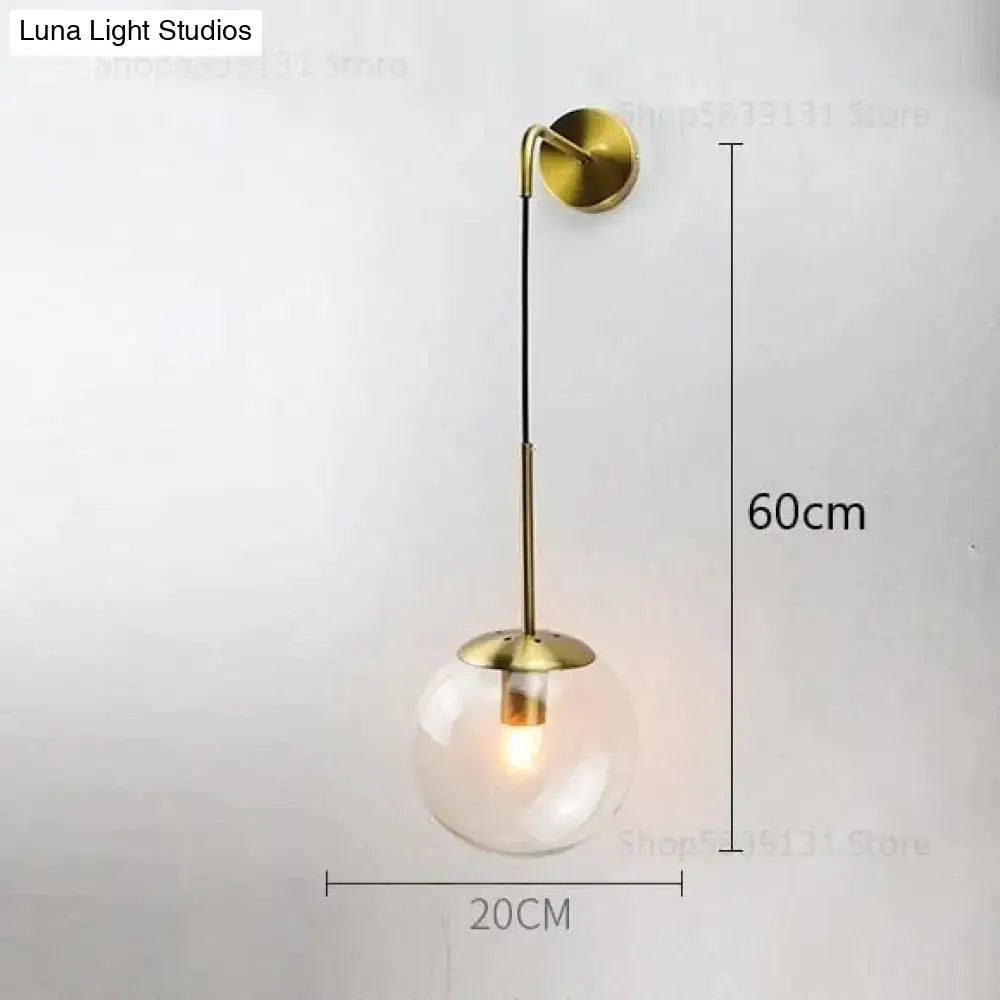 A sleek brass-finished retro modern glass wall lamp with clear globe and curved arm for bedroom lighting
