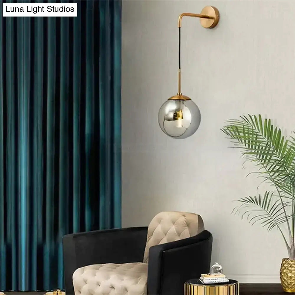 A sleek retro modern glass smoked globe wall lamp with a gold-toned metal fixture and black cord for bedroom lighting
