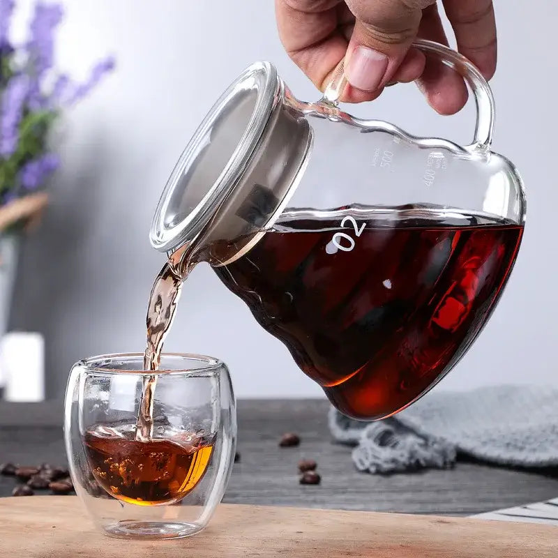 Clear glass coffee maker pot with silver lid pouring dark tea into double-walled glass cup for coffee brewing