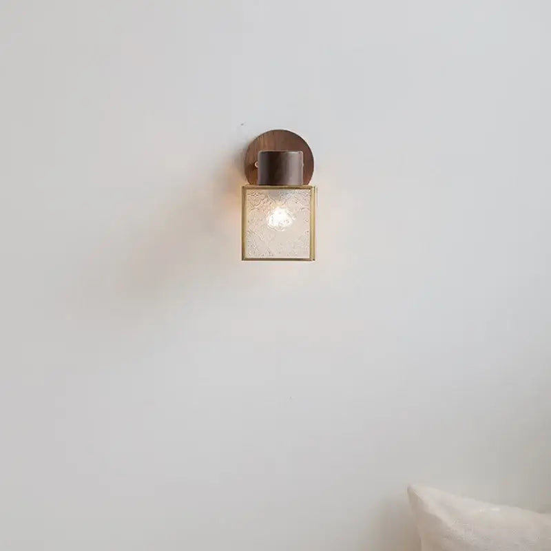 A modern wall sconce with walnut wood circular base, brass accents, and clear glass panels for bedroom lighting
