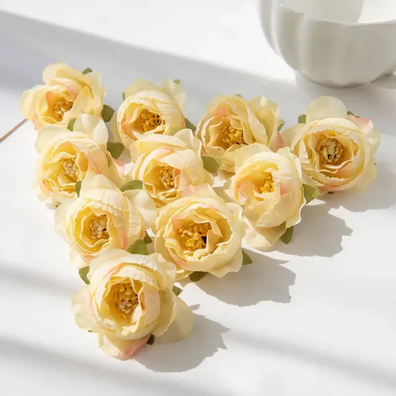 A cluster of cream-colored silk peony flowers with pink blush accents and green leaves on a white surface for decor