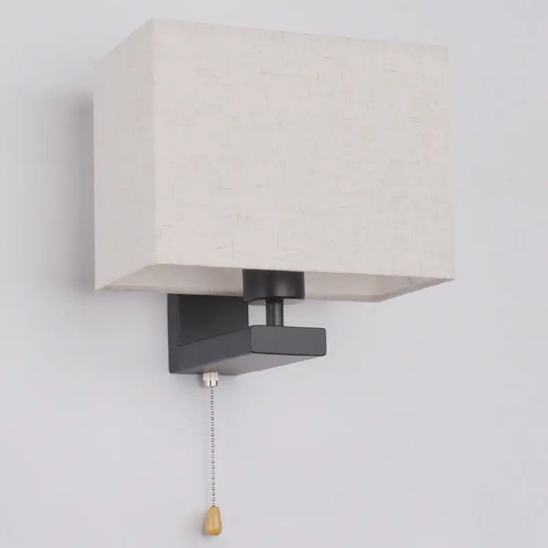 A modern black metal light fixture with an off-white fabric rectangle shade and wooden pull chain for bedroom use