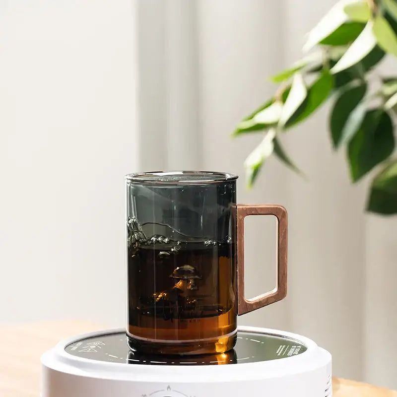 A sleek dark gray double walled glass tea cup with copper handle on a white electric heating base