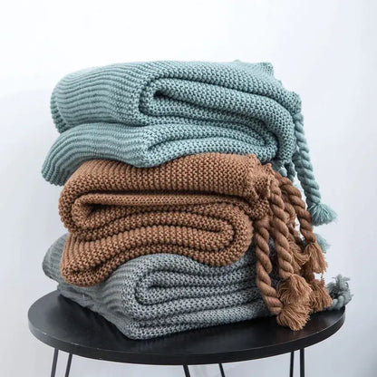 Stack of decorbites knit throw blankets in muted teal brown and gray with braided tassels for cozy decor
