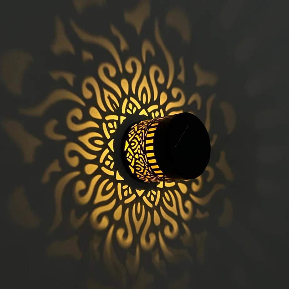 Black cylindrical solar-powered datura lantern with intricate golden cut-out patterns casting ornate shadows for outdoor decor