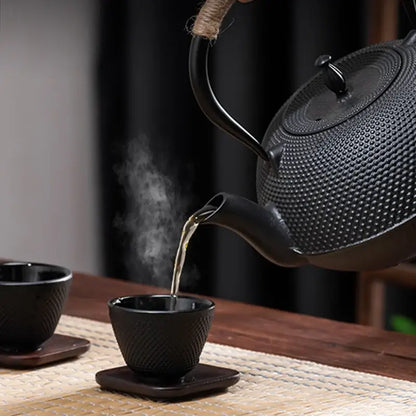 Textured black cast iron tea pot with woven handle pouring steaming tea into matching black cast iron tea cups