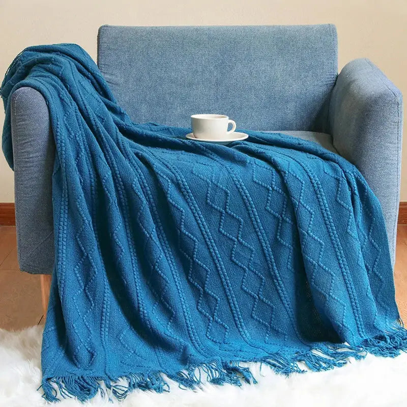 A cozy teal cable knit throw blanket with fringed edges draped over a blue armchair perfect for decorbites cozy knit decor