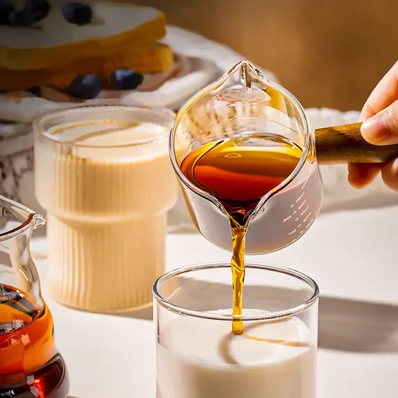 Transparent glass espresso measuring cup with wood handle filled with amber syrup being poured into a glass