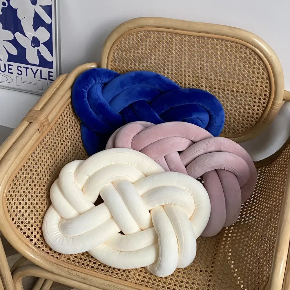 Three velvet knot throw pillows in royal blue soft pink and creamy white on a woven rattan chair for living room decor
