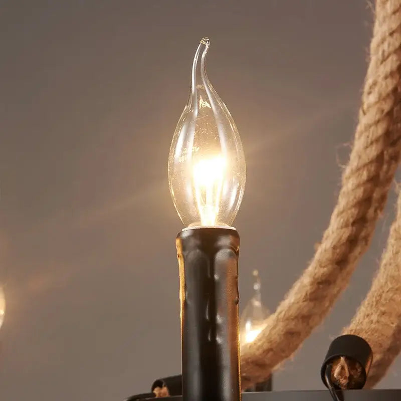 A glowing clear glass candle-style bulb with black wax-like base in a vintage black metal industrial hemp rope chandelier