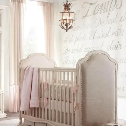Beige upholstered crib with arched headboard and nailhead trim alongside vintage decorbites grey metal cage lantern chandelier