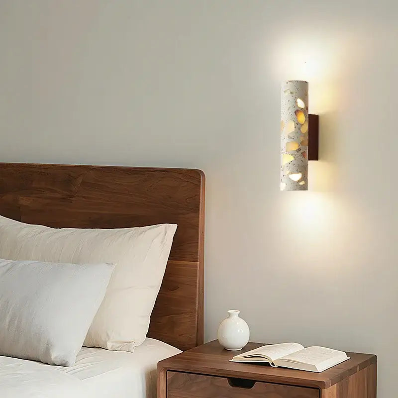 A cylindrical concrete wabi sabi terrazzo and wood led wall light with yellow and white pebbles mounted on dark wood bracket