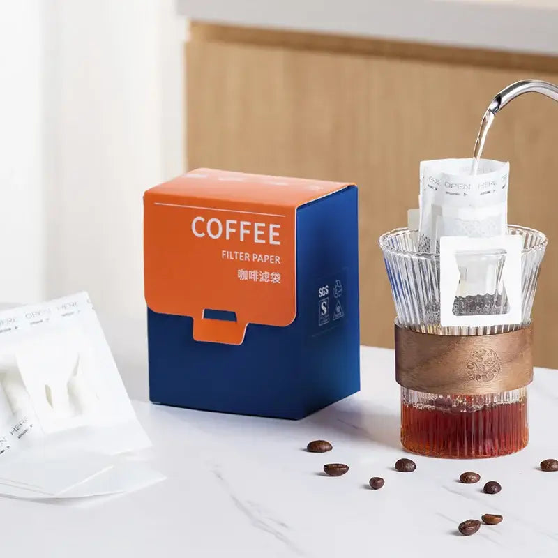 Orange and blue box of decorbites coffee filter bags next to a glass pour-over dripper with copper band for kitchen use