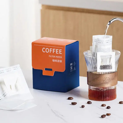 Orange and blue box of decorbites coffee filter bags next to a glass pour-over dripper with copper band for kitchen use