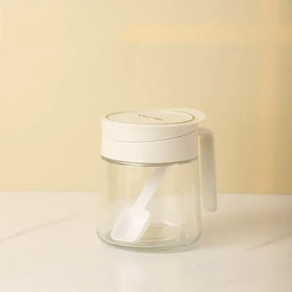 Clear glass seasoning jar with white lid handle and integrated spoon for kitchen spice shaking and storage